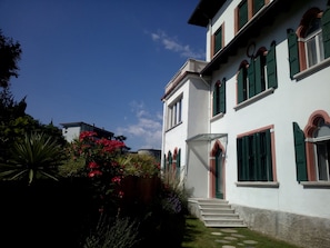 Edifici de dues plantes amb façana blanca i persianes verdes, que destaca una escala central i un jardí petit amb flors vermelles.
