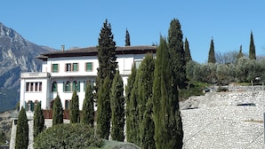 Un edificio blanco con contraventanas verdes, rodeado de altos cipreses y un camino de piedra.