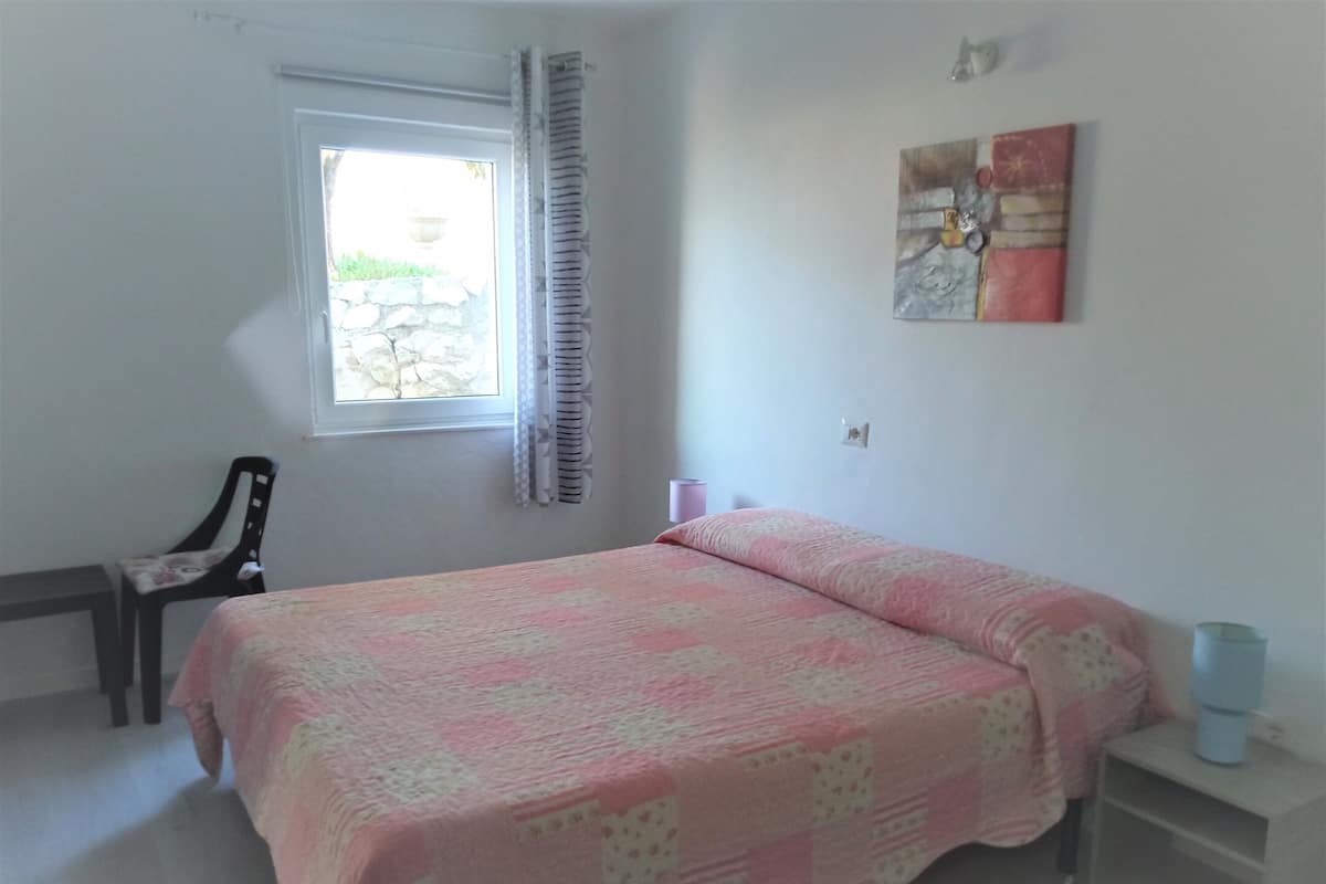 A bedroom with a bed, a chair, a window, a wall-mounted artwork, and a bedside table.
