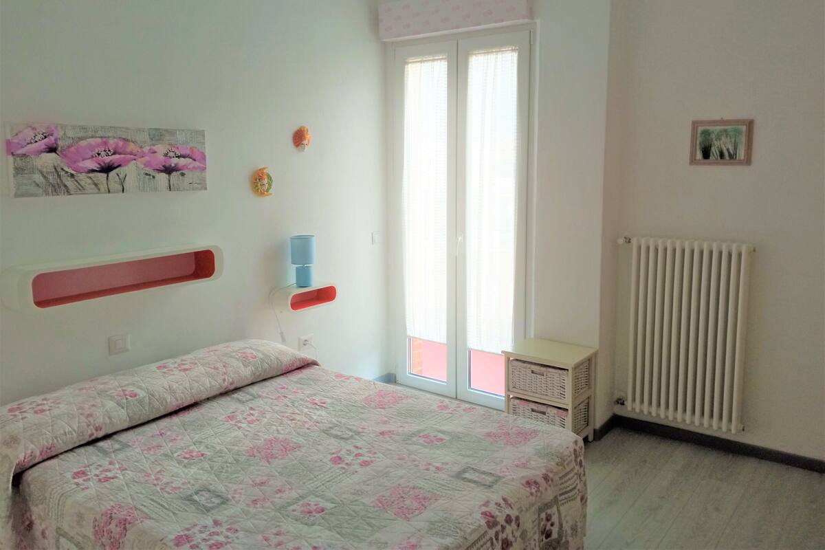 A bedroom with a bed, a window, a radiator, and a wall with a picture and a shelf.