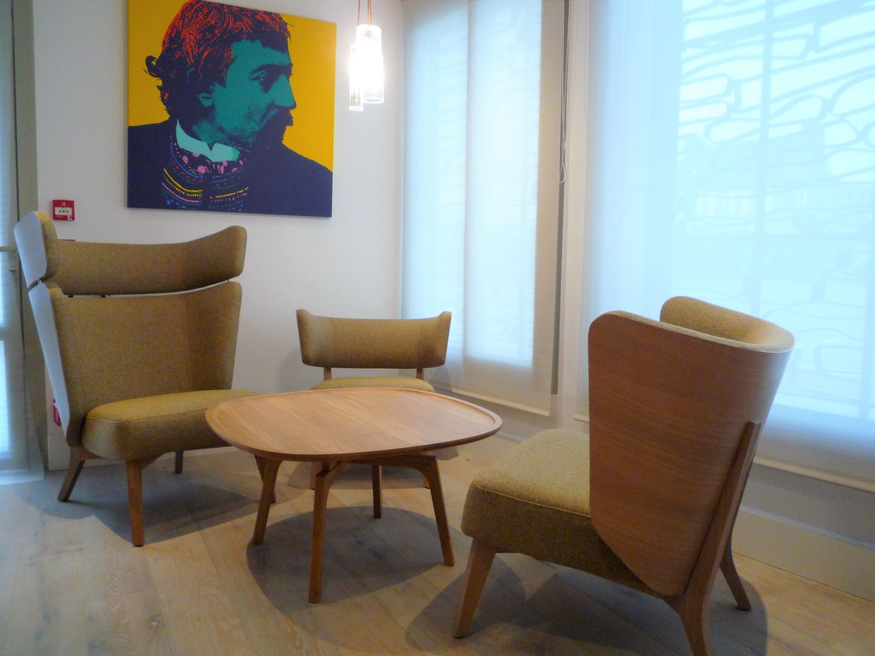 A room with three chairs and a small table, a painting on the wall, and a window with blinds.
