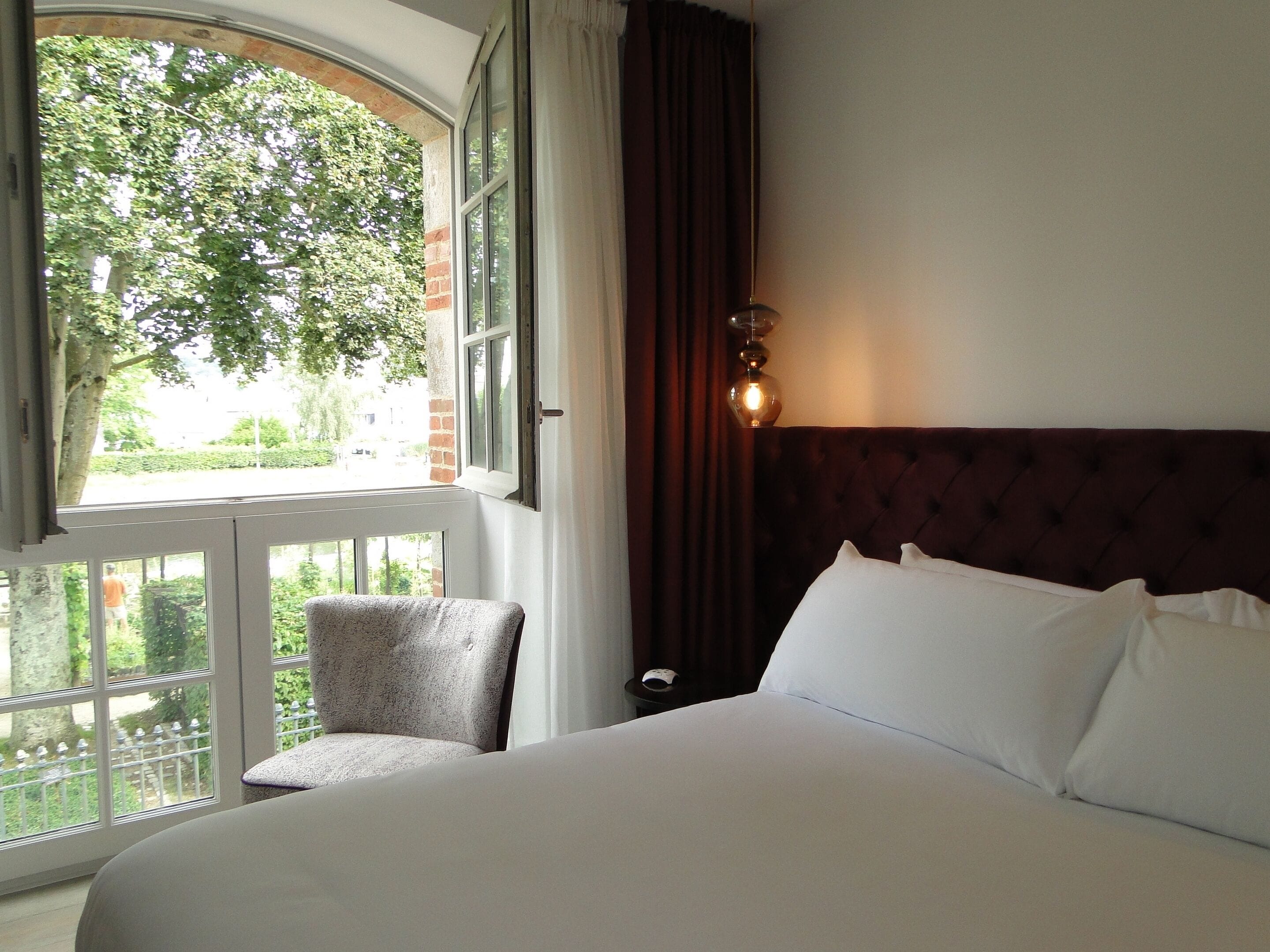 Une chambre d’hôtel avec un grand lit, une chaise, une fenêtre donnant sur un espace verdoyant et un luminaire fixé au mur.