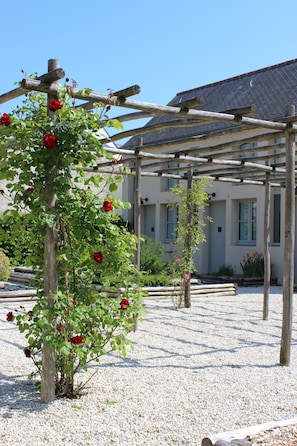 A pergola with climbing roses in a garden area with gravel ground, wooden posts, and a building with windows in the background. - Hôtel Ginkgo (Quimper)
