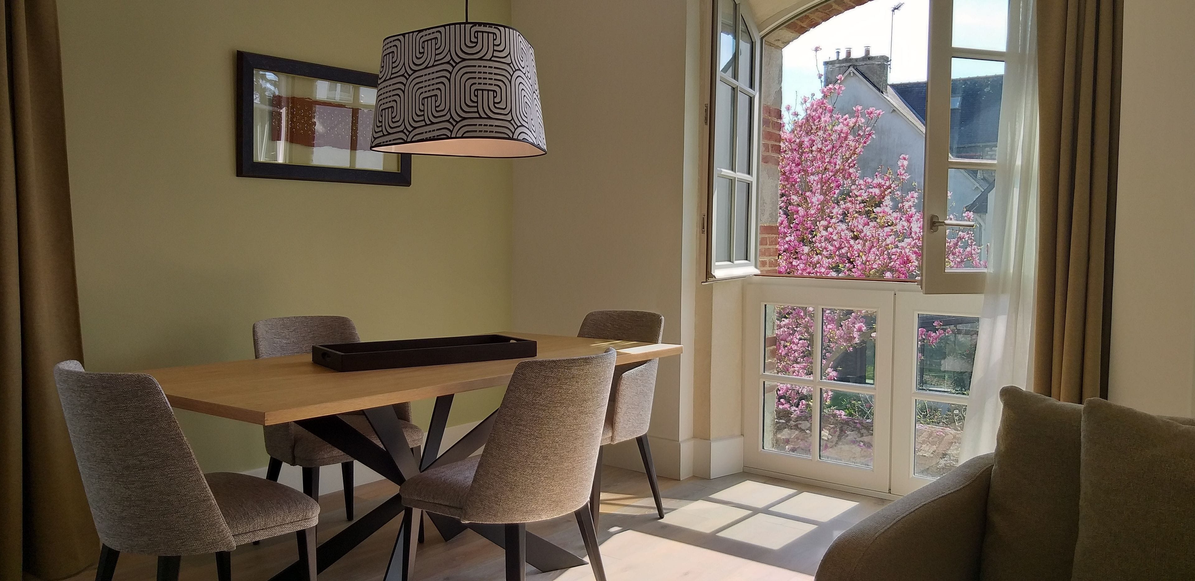 A dining area with a wooden table, six chairs, a black tray, a patterned pendant light, and a large window with pink flowers outside.