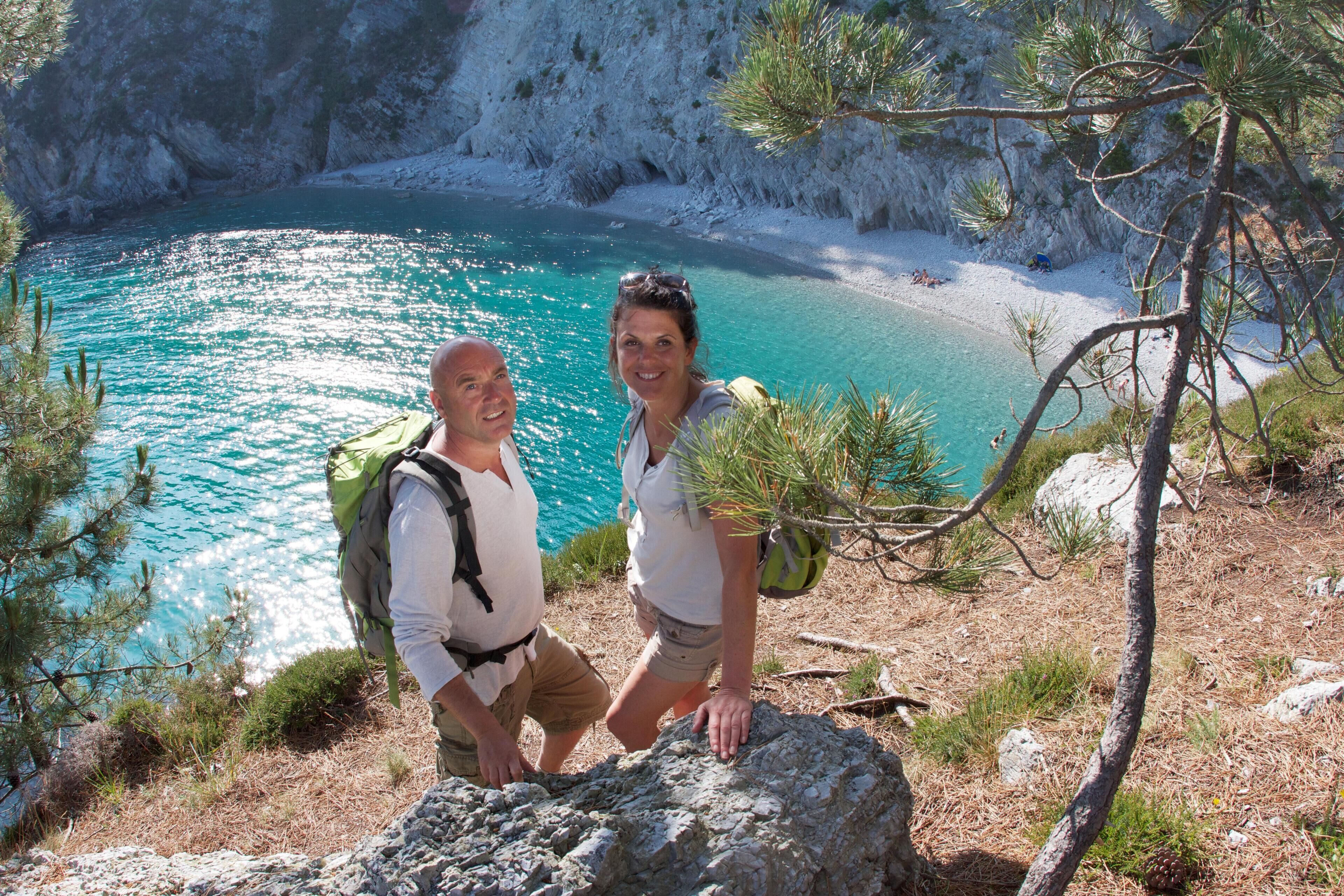 Twee personen staan op een rotsachtige richel met uitzicht op een turquoise zee en een wit zandstrand.