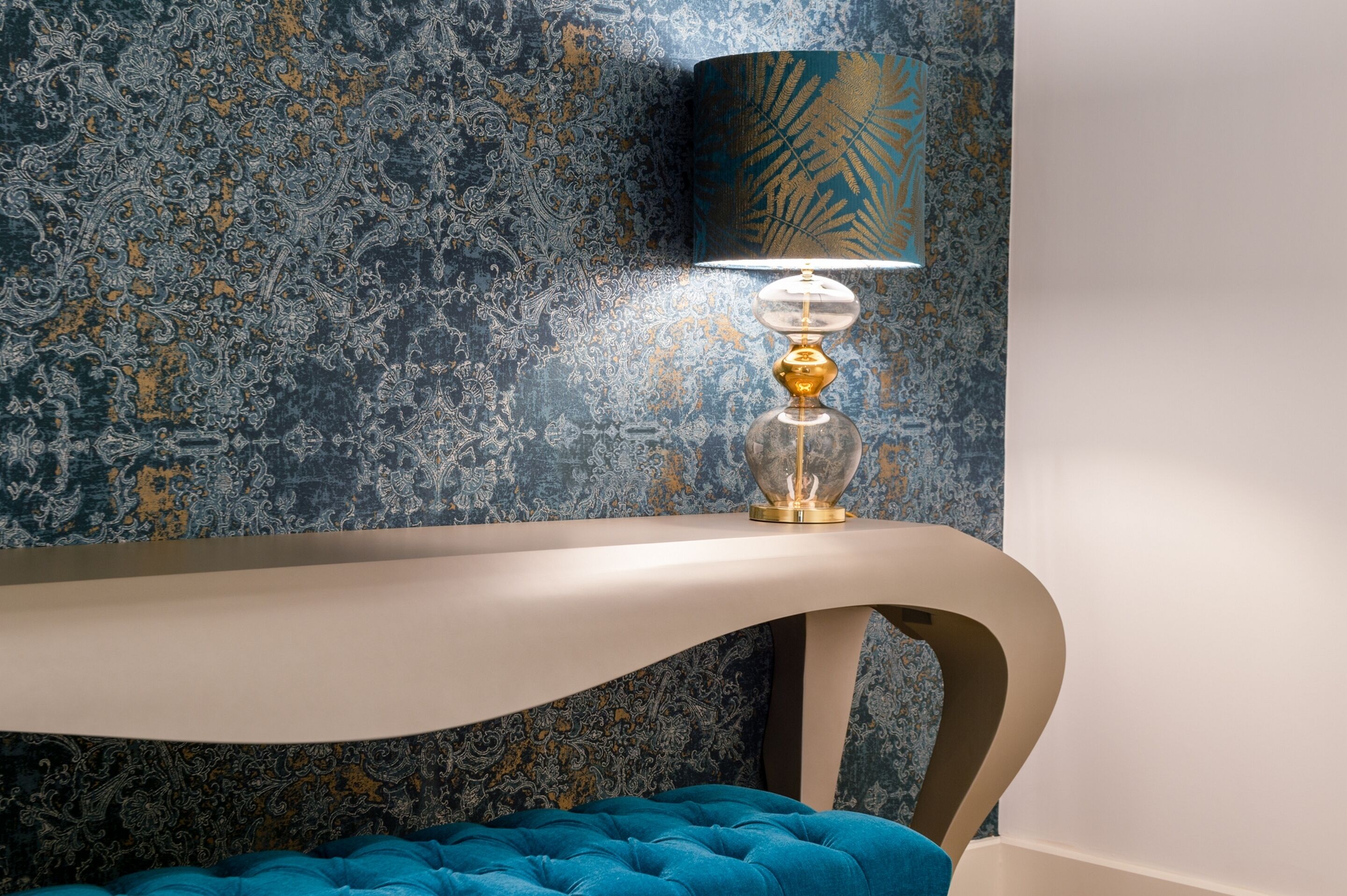 A modern room with a curved console table, a blue upholstered bench, and a wall-mounted lamp with a patterned shade.