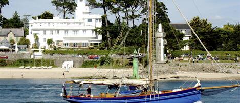 Een traditionele zeilboot op een kalm wateroppervlak met een strand en gebouwen op de achtergrond.