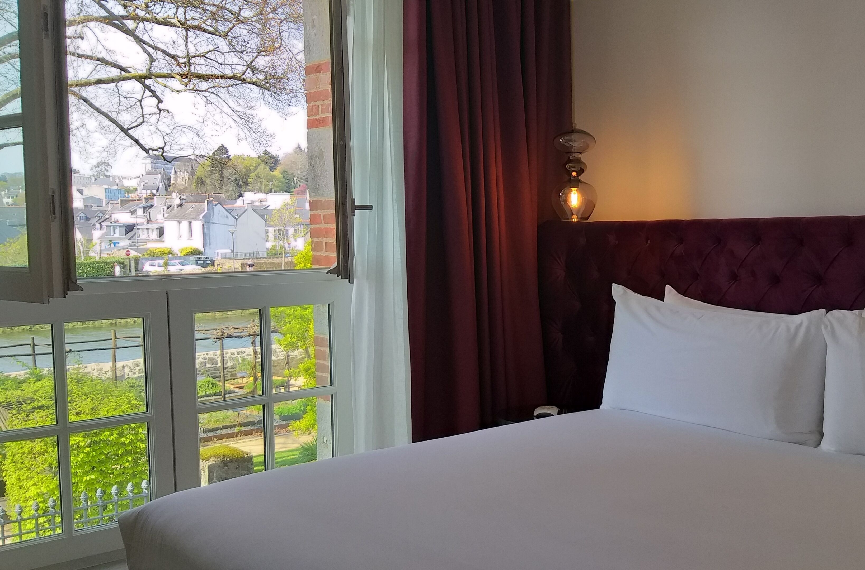 A hotel room with a large window, a bed with white linens, and a red headboard.