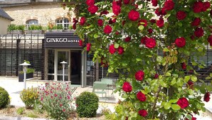 Entrada de hotel com portão de ferro forjado, placa indicando "GINKGO HOTEL" e um jardim com rosas vermelhas e outras plantas.