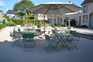 An outdoor patio with green metal chairs and tables, a large umbrella, and a garden area with benches and a pathway. - Hôtel Ginkgo (Quimper)