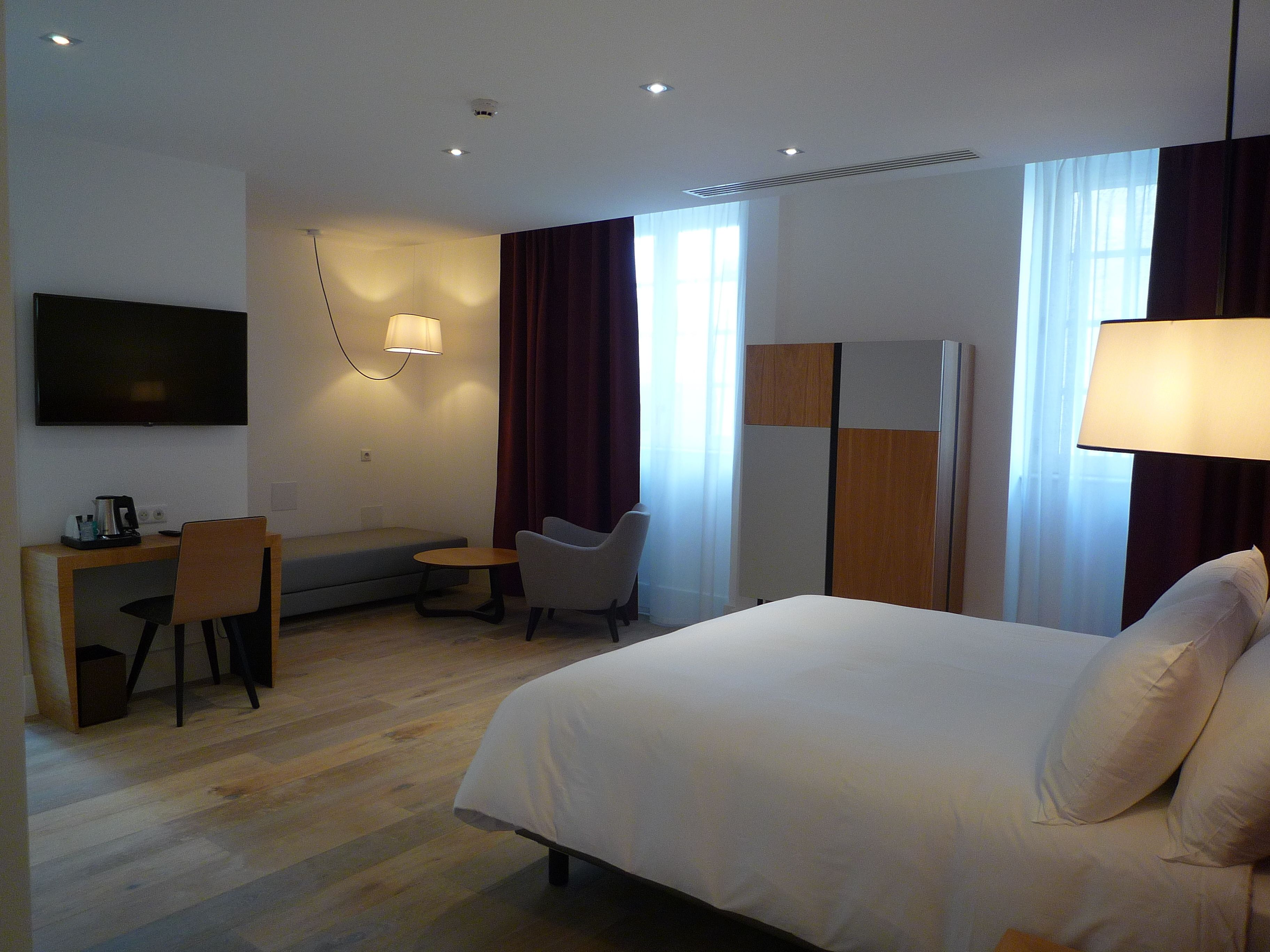 A hotel room with a bed, a desk with a chair, a TV, and a lamp.
