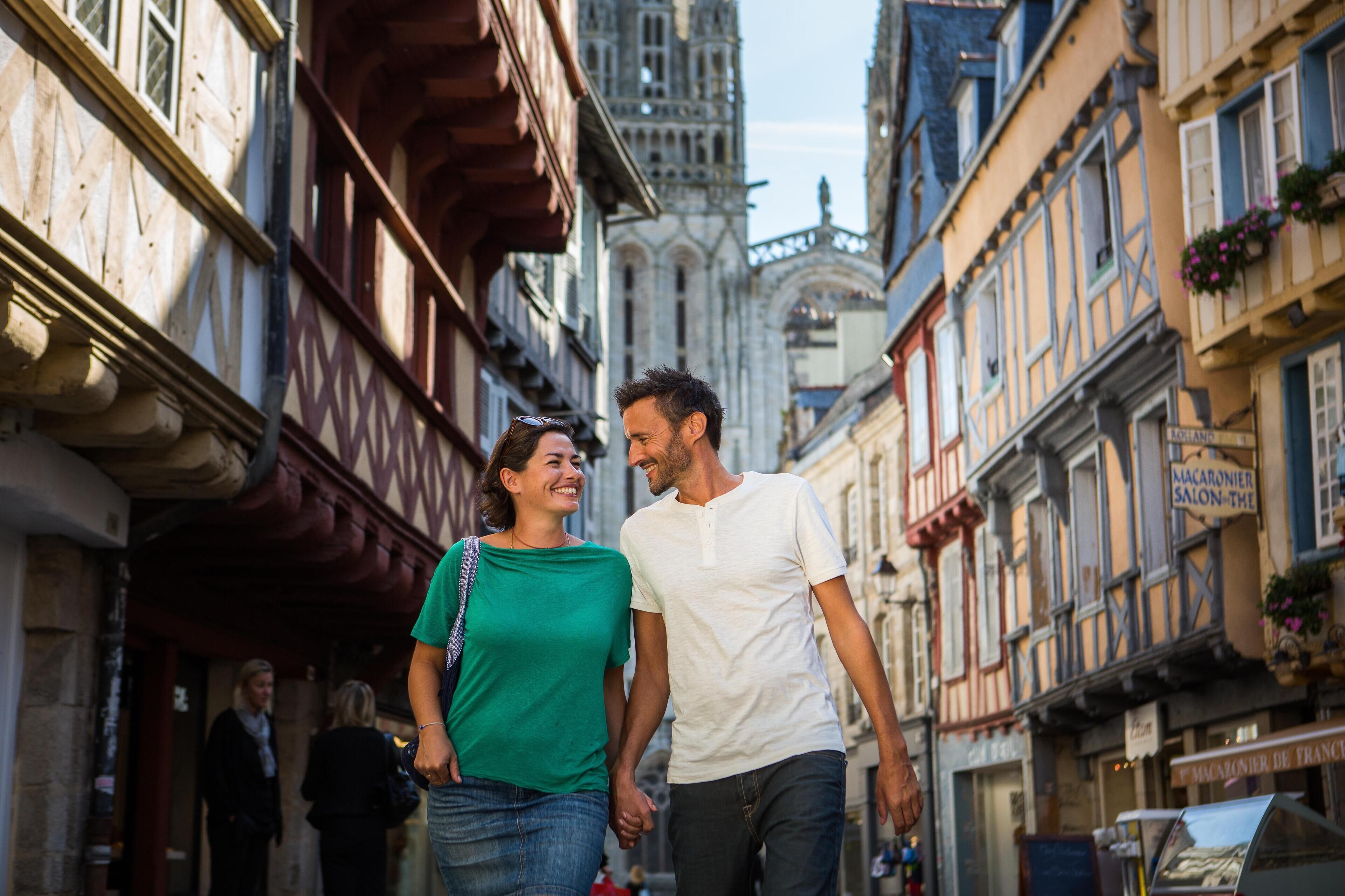 A couple walking on a narrow street with historic buildings, including a cathedral in the background.