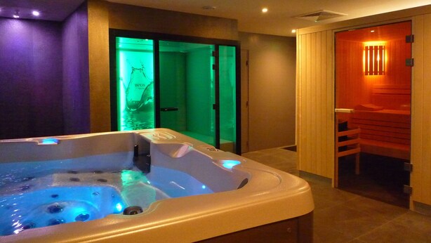 Um espaço de spa com hidromassagem, uma porta de madeira que dá para uma sauna, e uma porta de vidro iluminada em tons de verde.