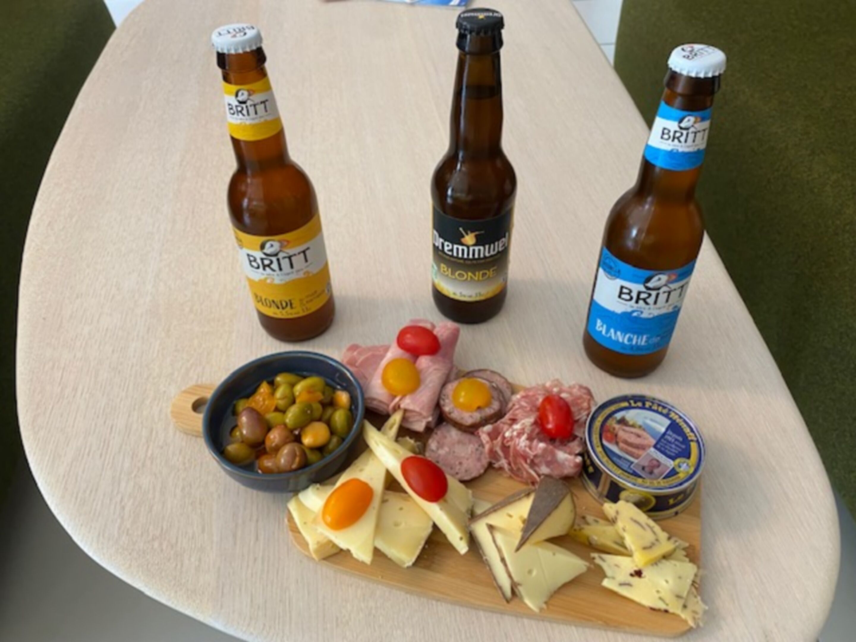 Een houten tafel met drie bierflesjes, een schaal olijven, diverse kazen en vleessoorten.