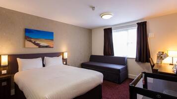 A hotel room with a bed, a seating area, a window with curtains, and a painting on the wall.