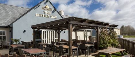 An outdoor seating area with tables and chairs, a wooden fence, and a building with "The Sessile Oak" sign.