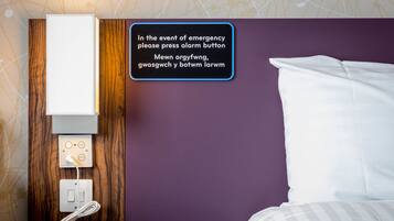 Hotelkamer met een bed, een bedieningsknop aan de muur, en een bedieningspaneel met een stopcontact en een telefoon.