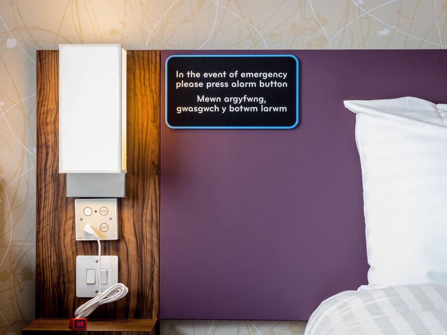 A hotel room with a bed, a wall-mounted alarm button, and a control panel with a power outlet and a phone.