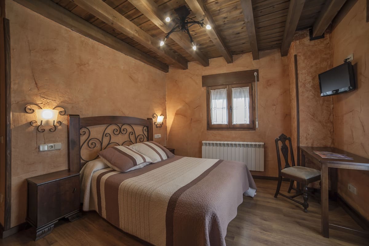 Un dormitorio con techo de madera, una cama con ropa de cama a rayas, una mesita de noche, un escritorio, una silla y un televisor en la pared.