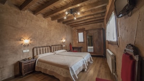 A rustic bedroom with wooden beams, two beds, a nightstand, a red chair, and a television. - La Casona del Herrero (Navaleno)