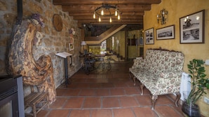A rustic interior with a stone wall, wooden beams, a patterned sofa, a table with a lamp, and framed pictures on the wall. - La Casona del Herrero (Navaleno)