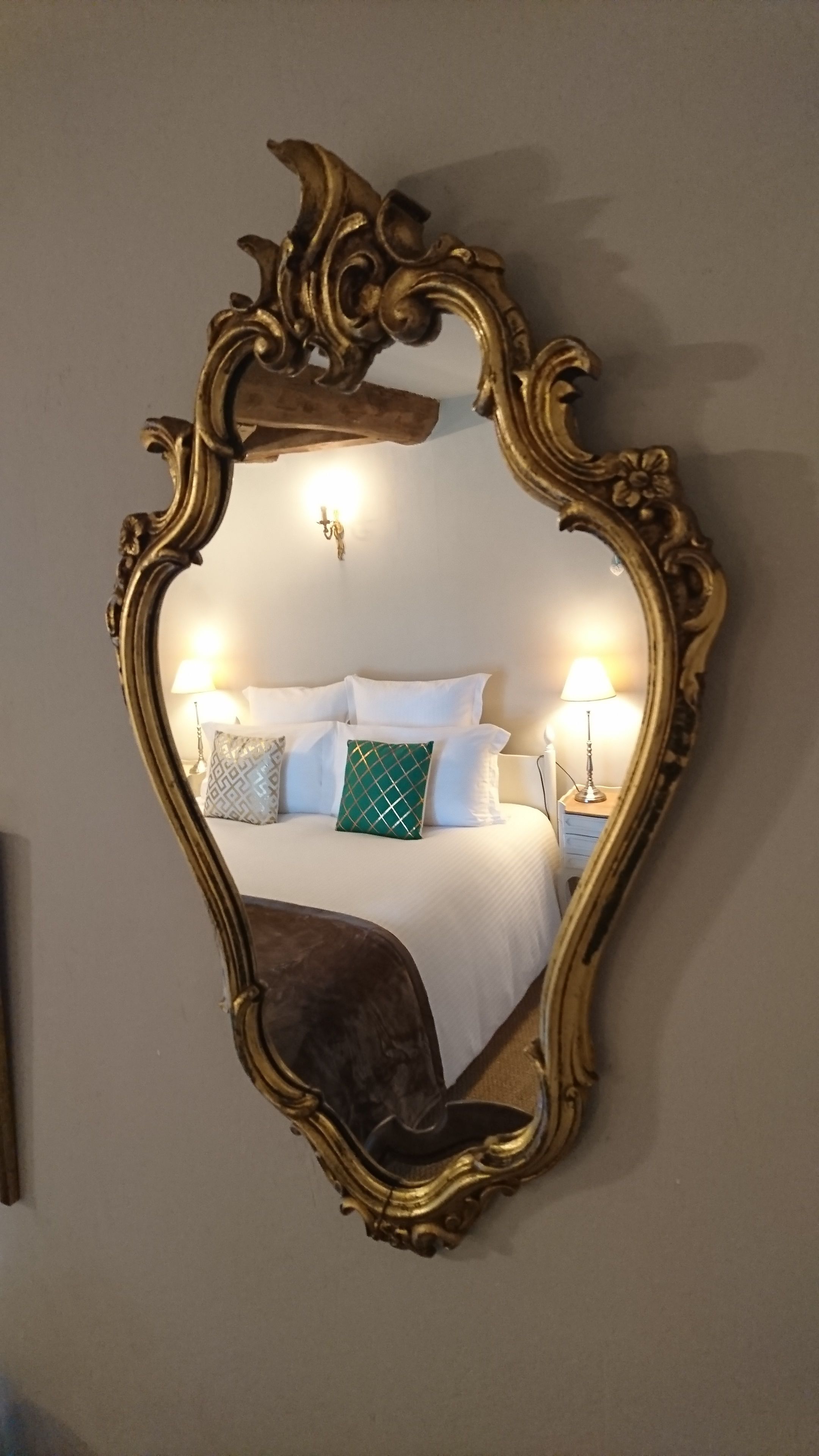 A mirror with an ornate gold frame reflecting a bed with white bedding and two lamps.