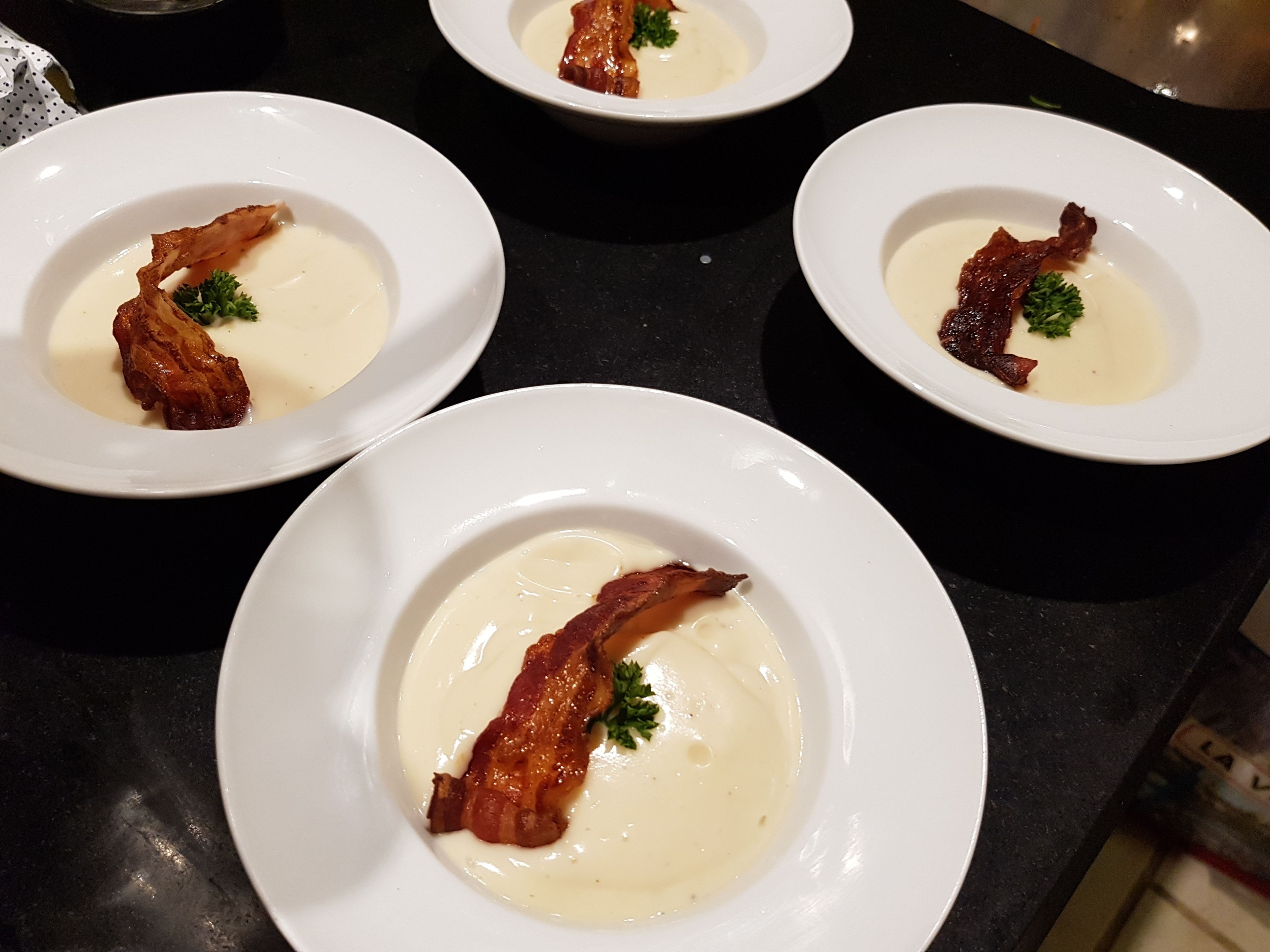 Three white plates with a creamy soup and crispy bacon garnish on a black countertop.