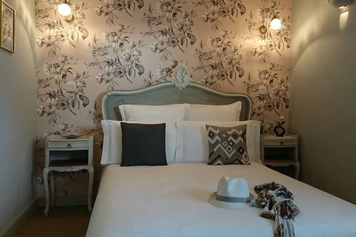 En pænt redt seng med puder og en hat, et natbord med en lampe, og blomstret tapet.