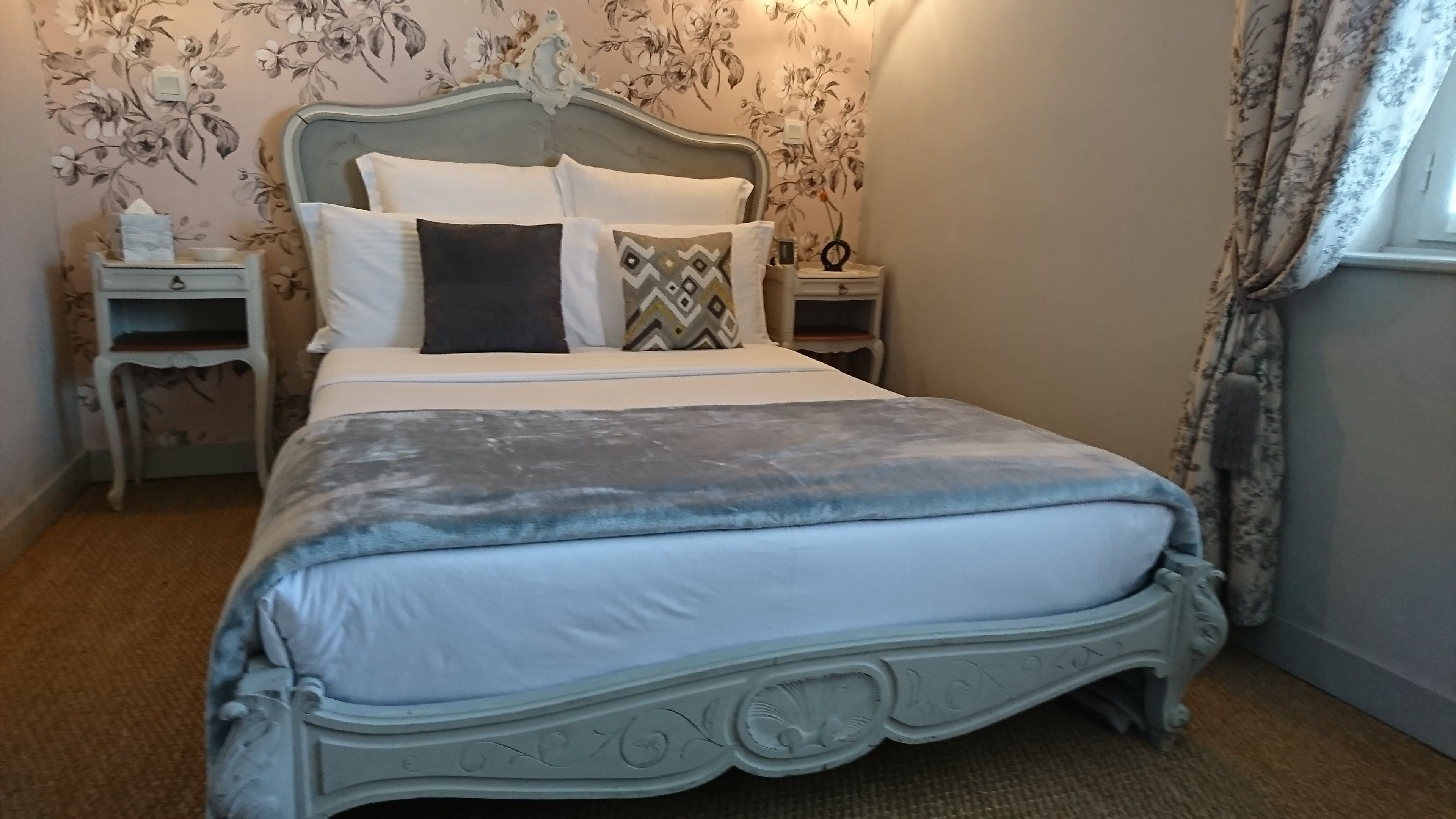 A neatly made bed with a grey blanket and decorative pillows, a wooden headboard, and floral wallpaper.