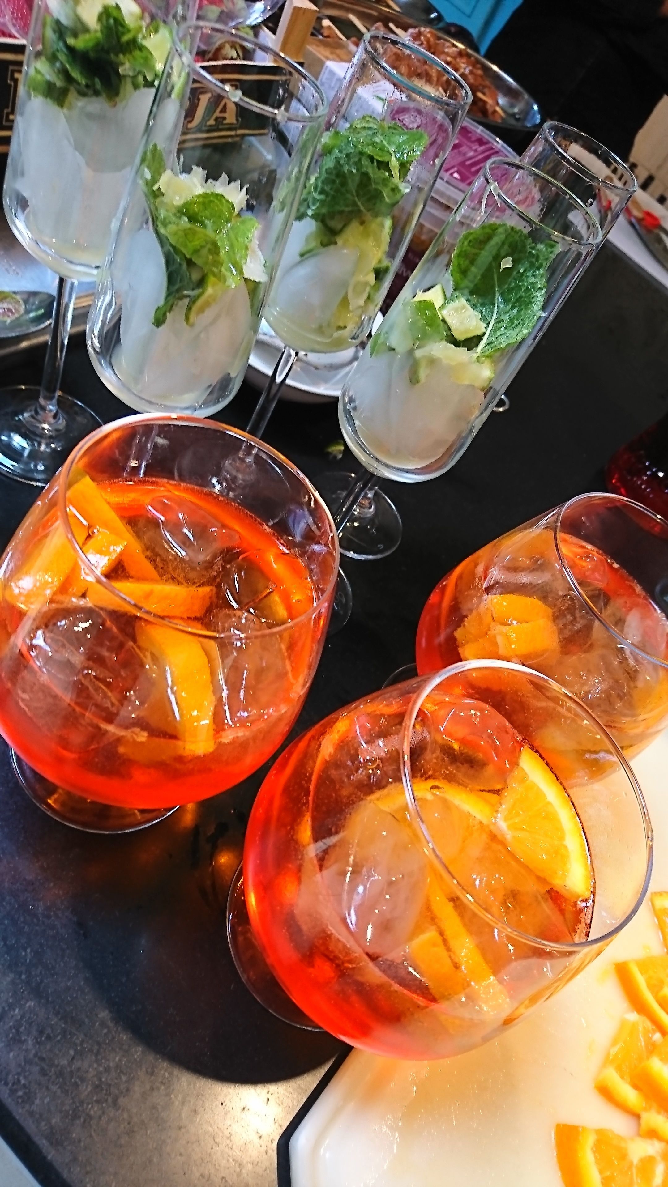 Una zona de bar con varios vasos de cócteles, algunos con hielo y guarniciones, y una bandeja con más bebidas y adornos.