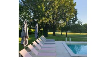 Un área junto a la piscina con sillones de descanso y sombrillas, rodeada de árboles y un cielo despejado.