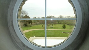 Een rond raam met uitzicht op een gazon, bomen en een verhard pad.