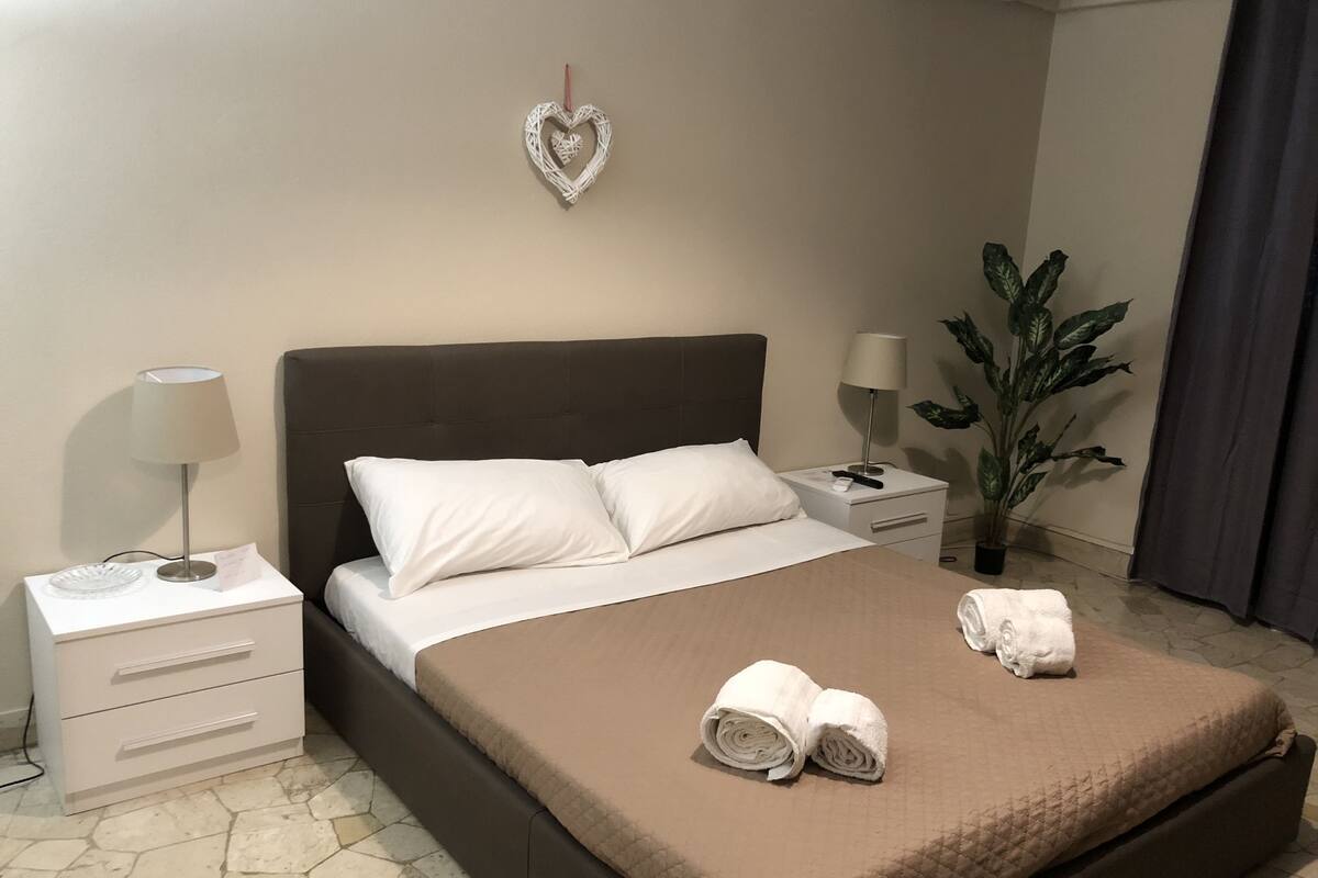 Cama bem arrumada com travesseiros brancos, manta marrom e duas toalhas enroladas. Há uma mesa de cabeceira branca com uma luminária, uma pequena planta em vaso e um coração decorativo na parede.