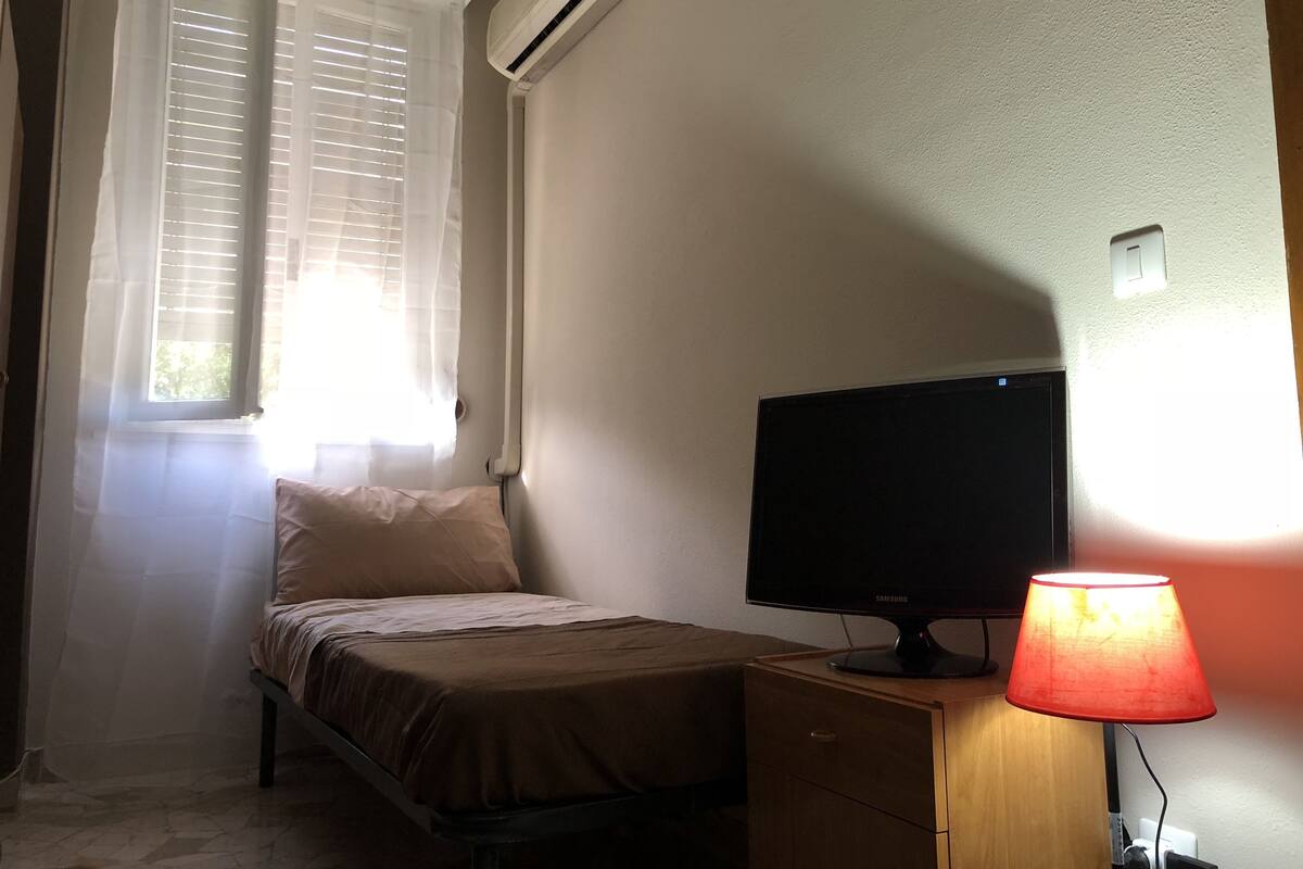 Quarto pequeno com cama, televisão, abajur e ar condicionado.
