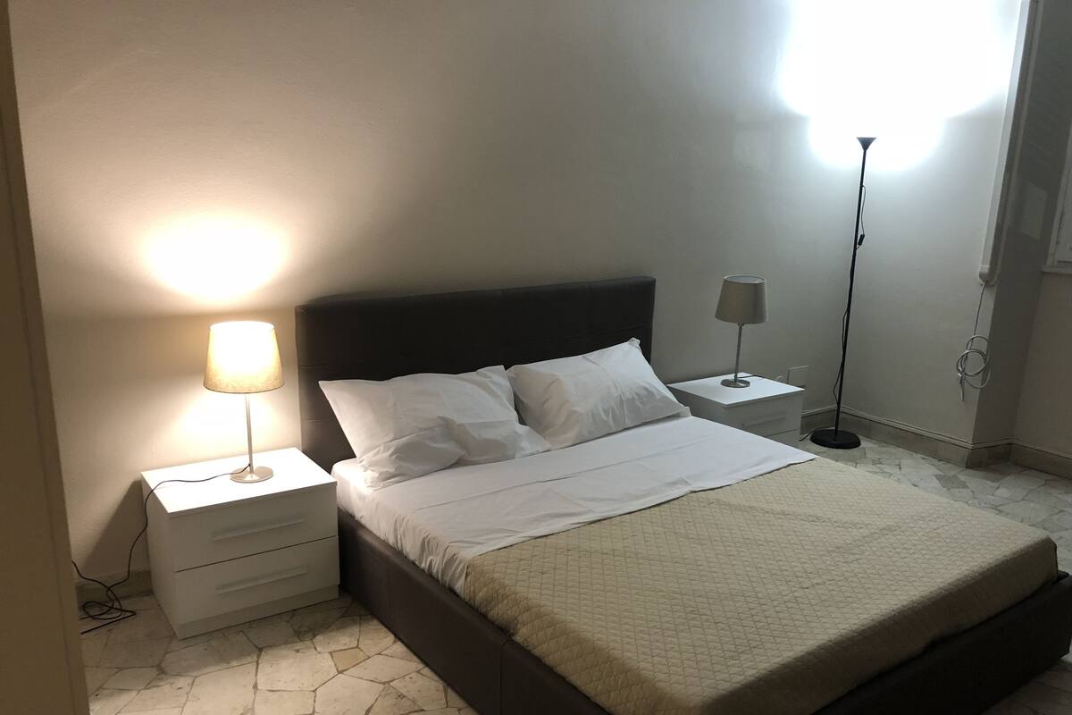 Cama bem arrumada com roupa de cama branca, cabeceira escura e mesa de cabeceira com abajur. O quarto tem piso de azulejos e parede lisa.