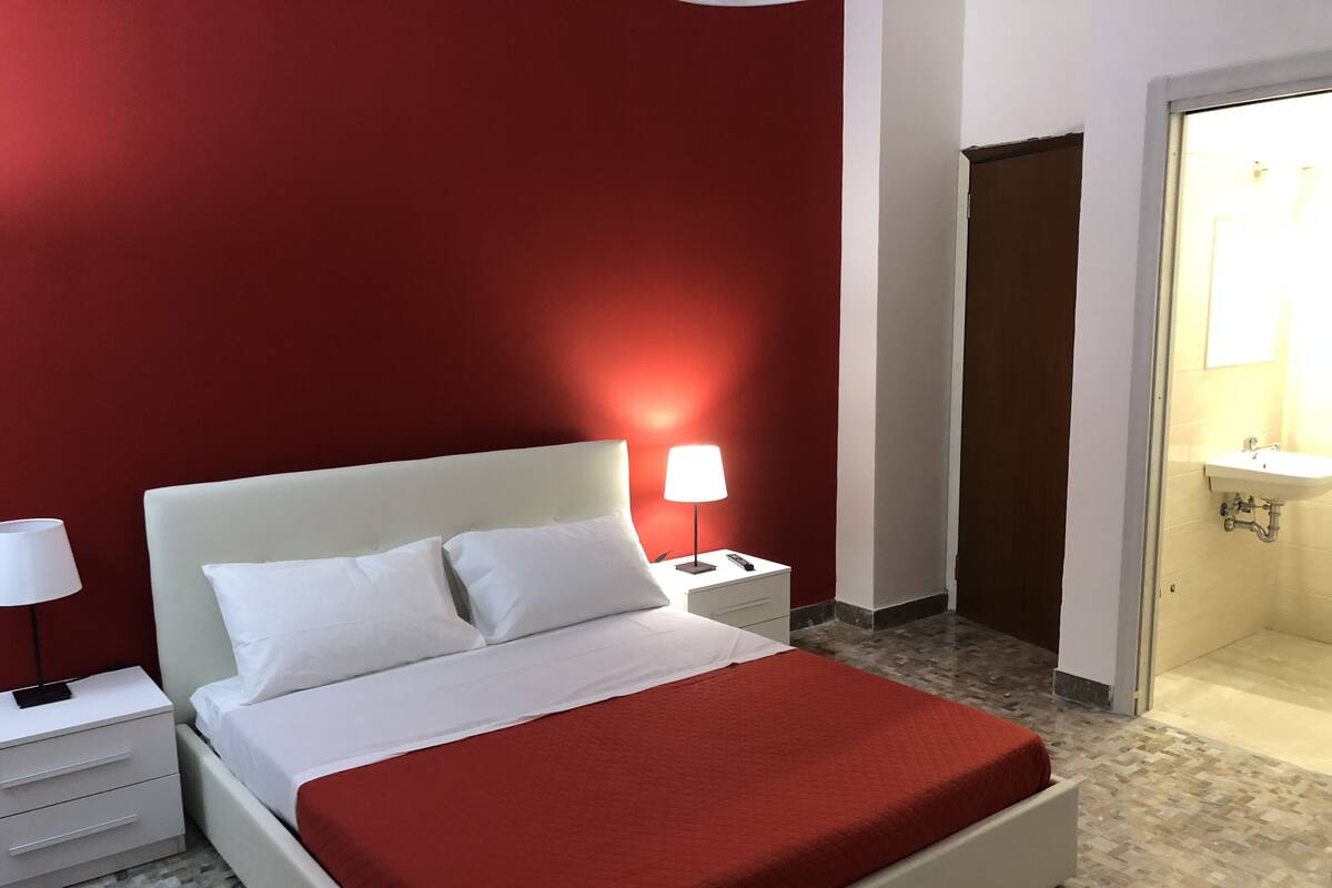 Quarto de hotel com cama, mesa de cabeceira, abajur e banheiro visível através de uma porta aberta.