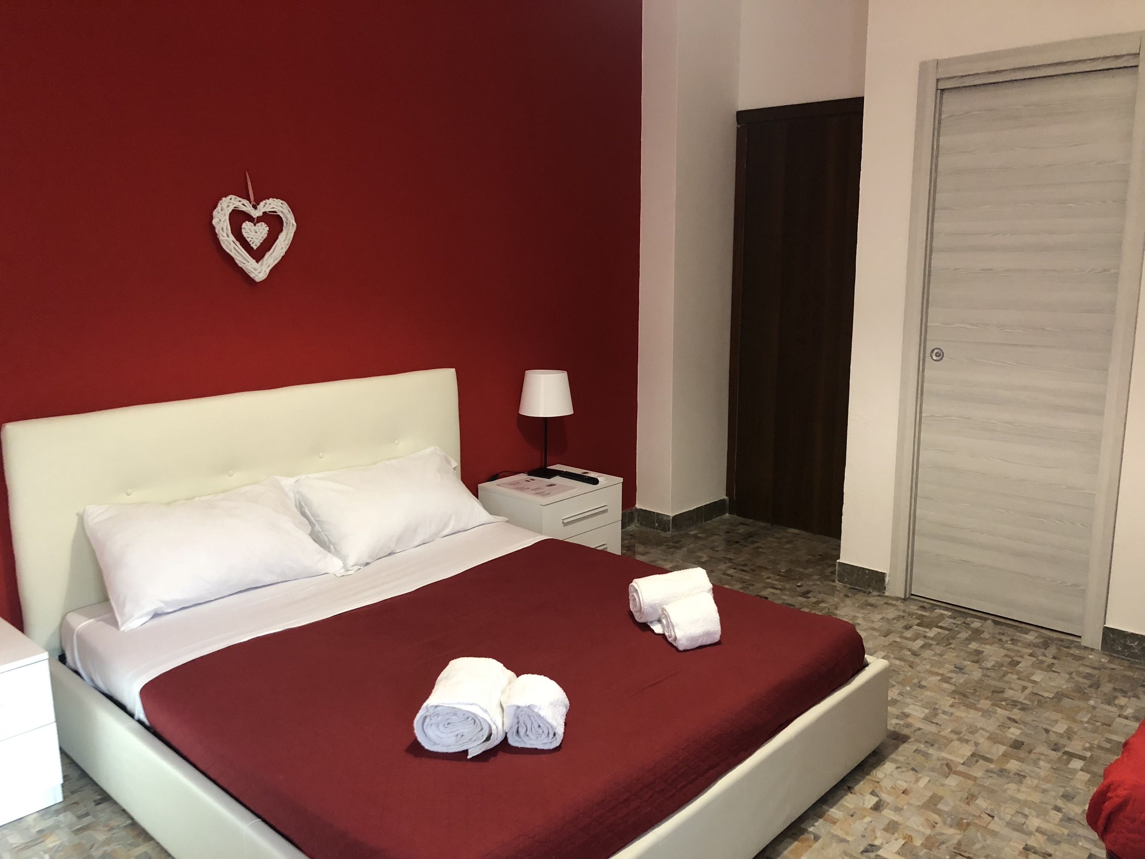 A neatly made bed with white linens and a red bedspread, a white nightstand with a lamp, a heart-shaped decoration on the wall, and a closed door in the background.