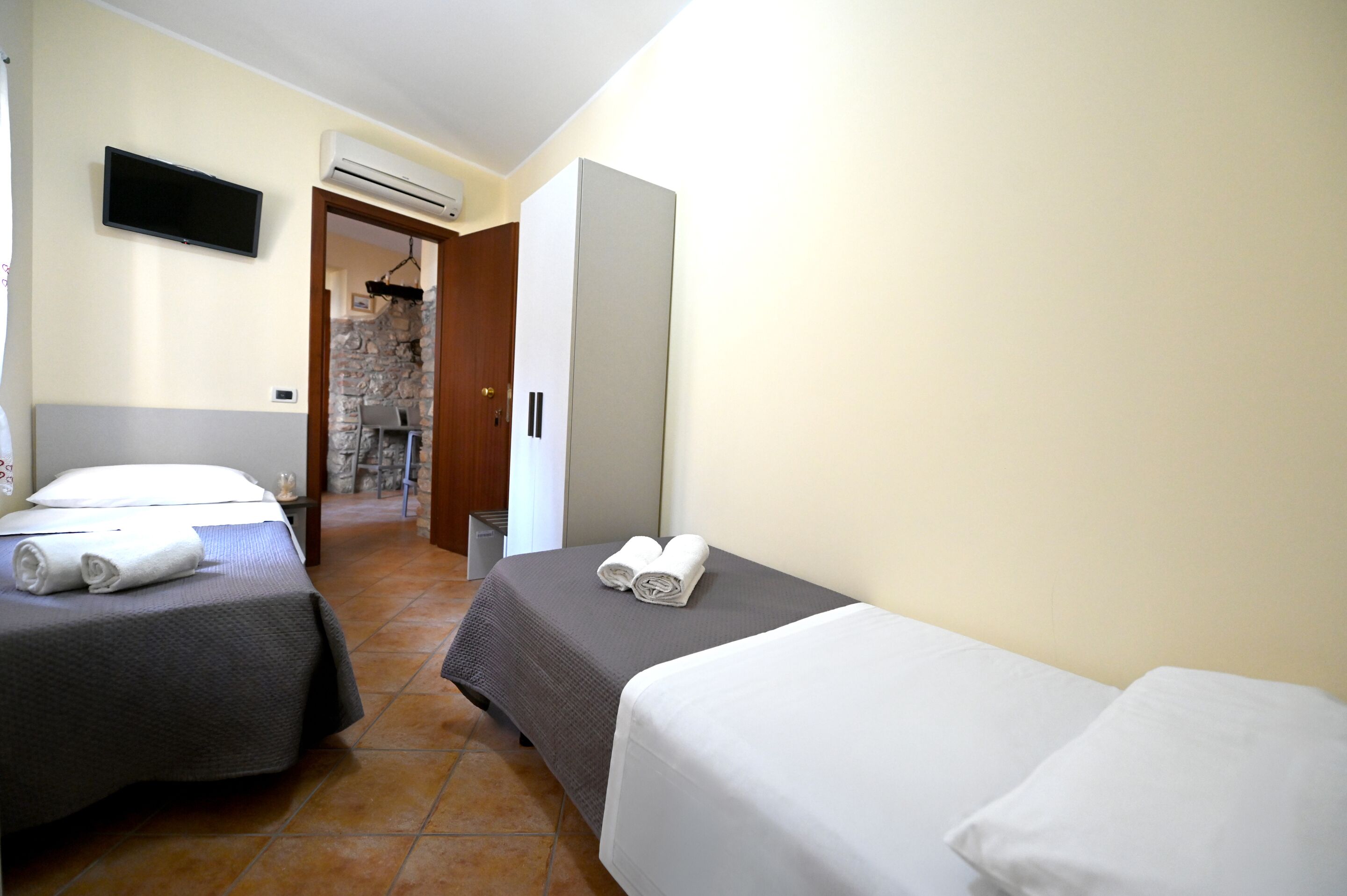 A hotel room with two beds, a TV, an air conditioning unit, and a stone wall visible through an open door.