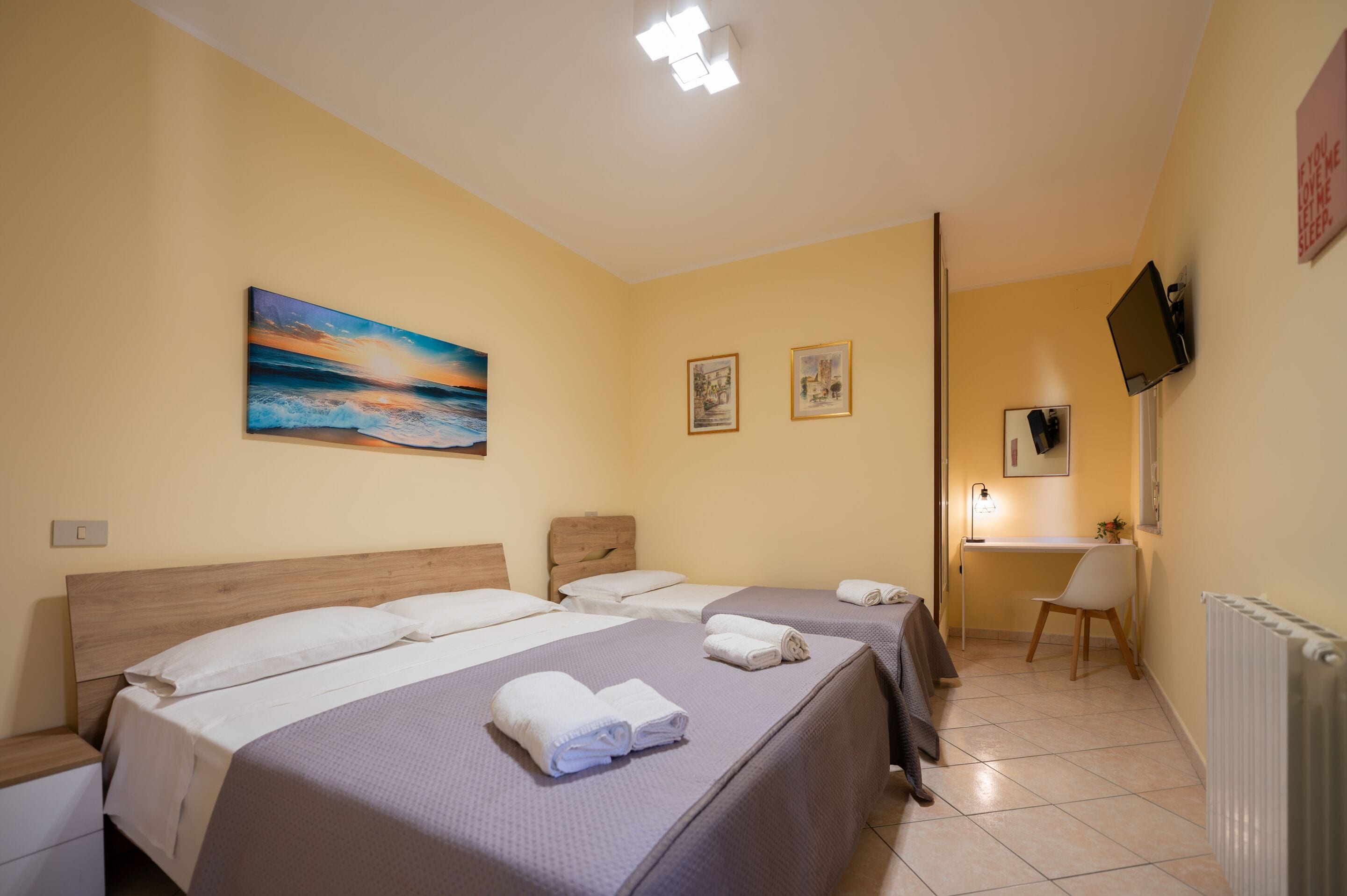A hotel room with three beds, a desk, a chair, a TV, and a painting on the wall.
