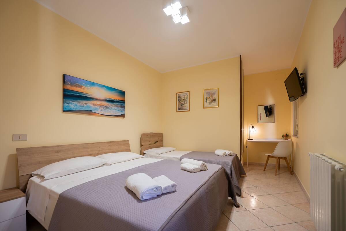 A hotel room with three beds, a desk, a chair, a TV, and a painting on the wall.