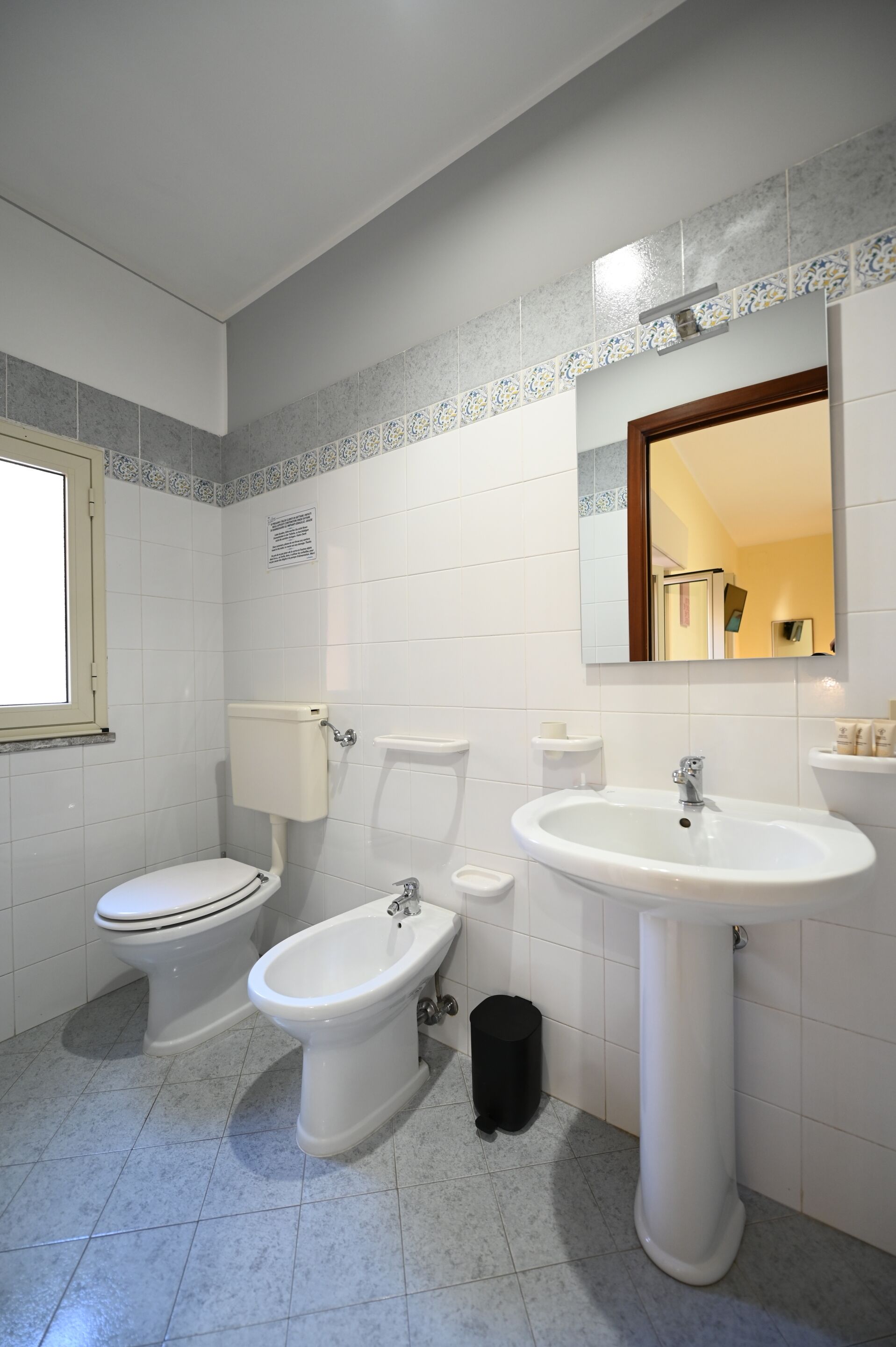 A bathroom with a toilet, bidet, sink, and a trash bin.