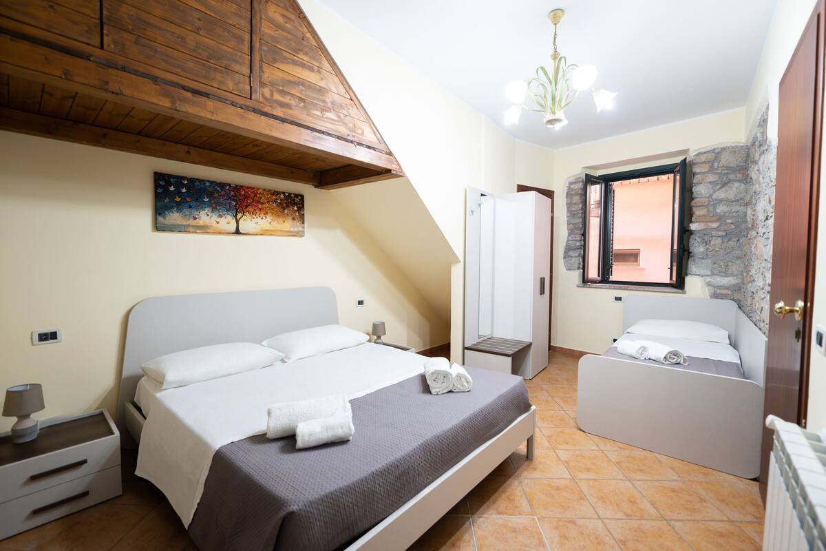 A bedroom with a bed, bedside tables, a nightstand, a painting, a wardrobe, and a window with a stone wall.