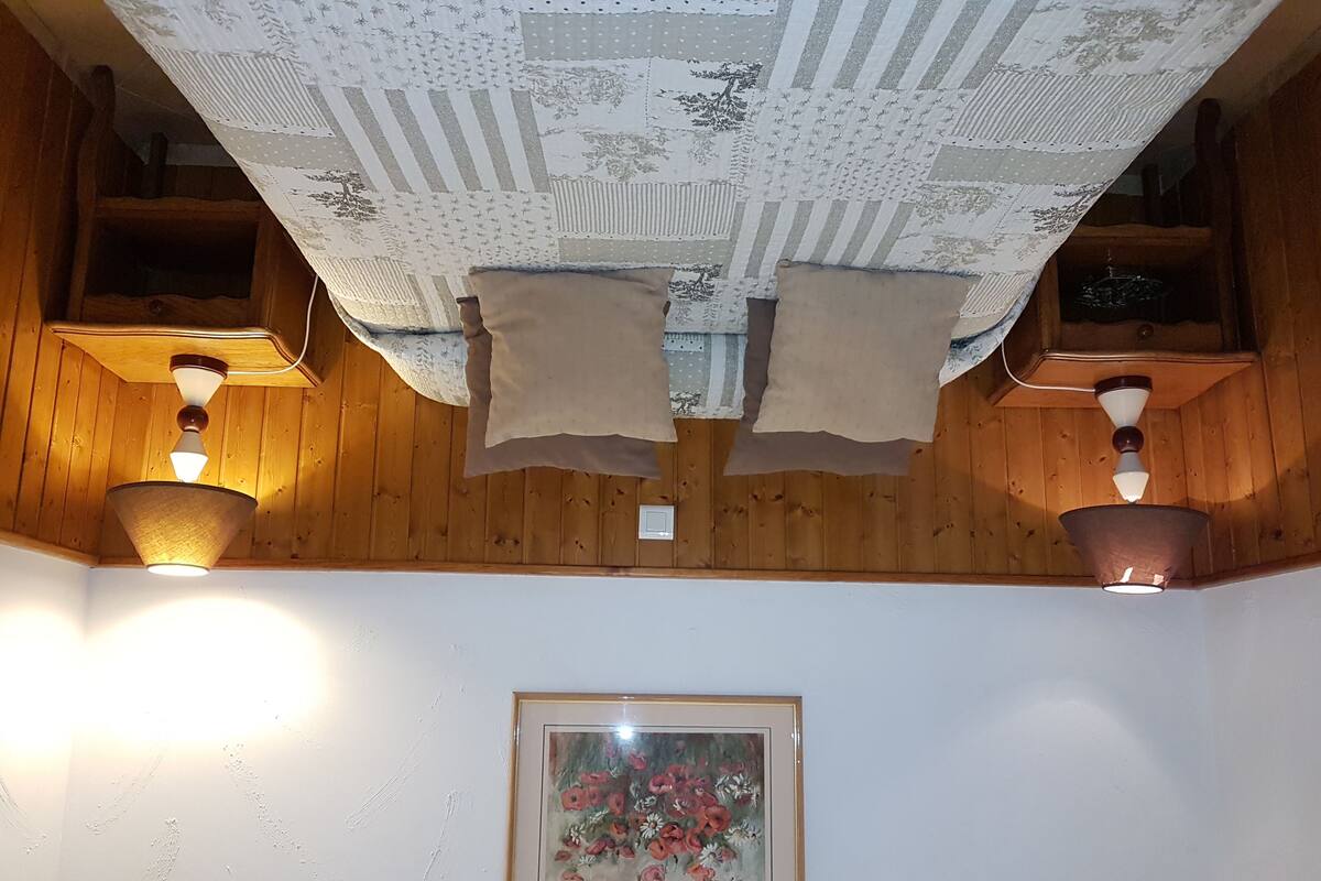 Huone, jonka seinät ovat puuta, katossa on kukka-aiheinen kuvio, siinä on kaksi seinälle asennettua lamppua ja kehystetty kukka-aiheinen maalaus.
