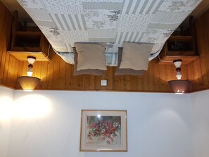 A room with wooden walls, a ceiling with a floral pattern, two wall-mounted lamps, and a framed floral painting. - Maison Amets (Sare)