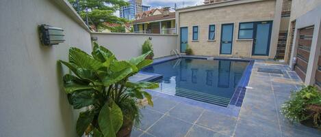 A swimming pool with a tiled deck, surrounded by buildings and potted plants.
