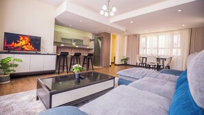 Un salon moderne avec un téléviseur à écran plat, une table basse et un canapé. On aperçoit à l’arrière-plan un coin cuisine avec une table à manger et des chaises.