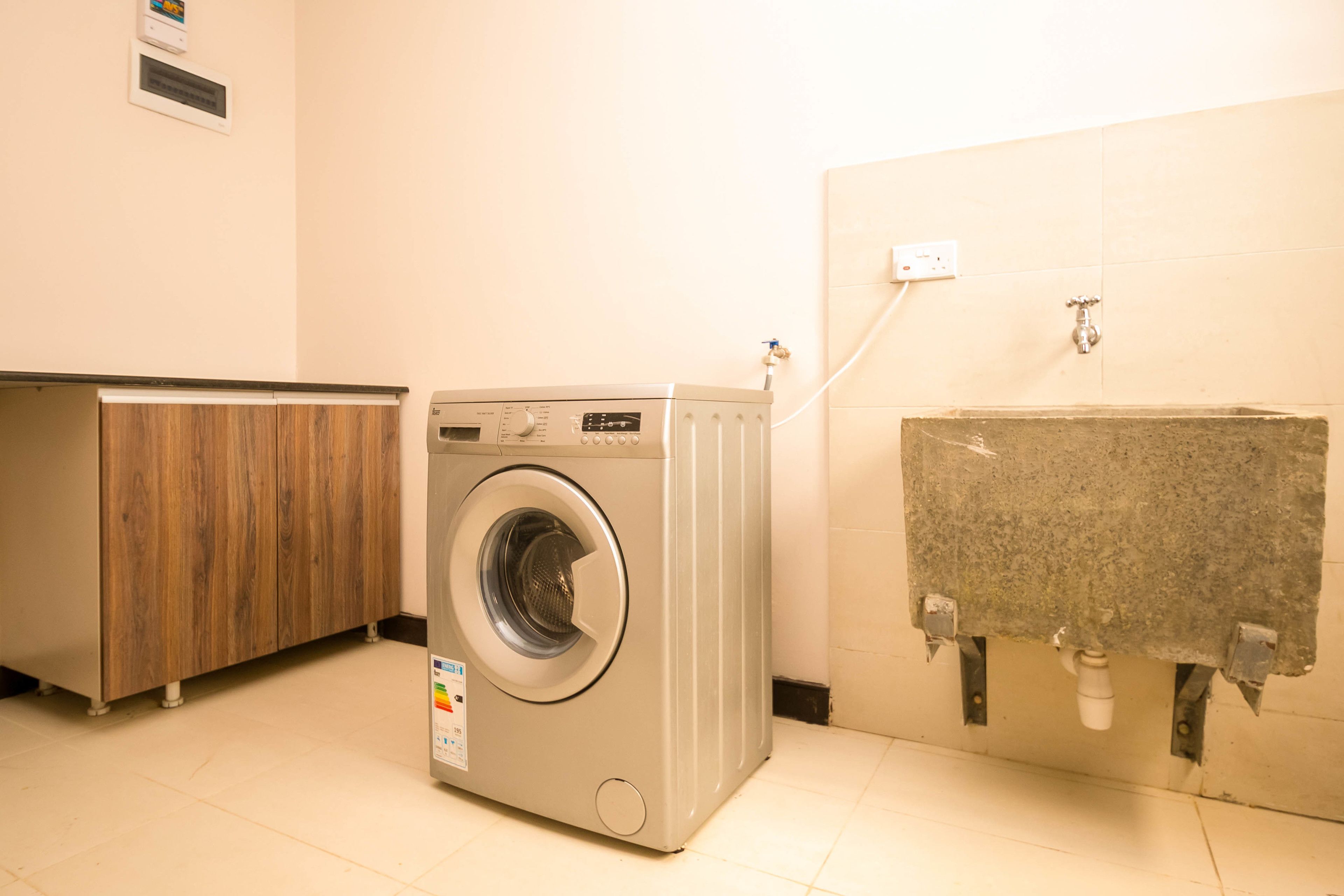 Une petite buanderie moderne, dotée d’un lave-linge à chargement frontal, d’un meuble en bois et d’un évier dont la tuyauterie est visible.