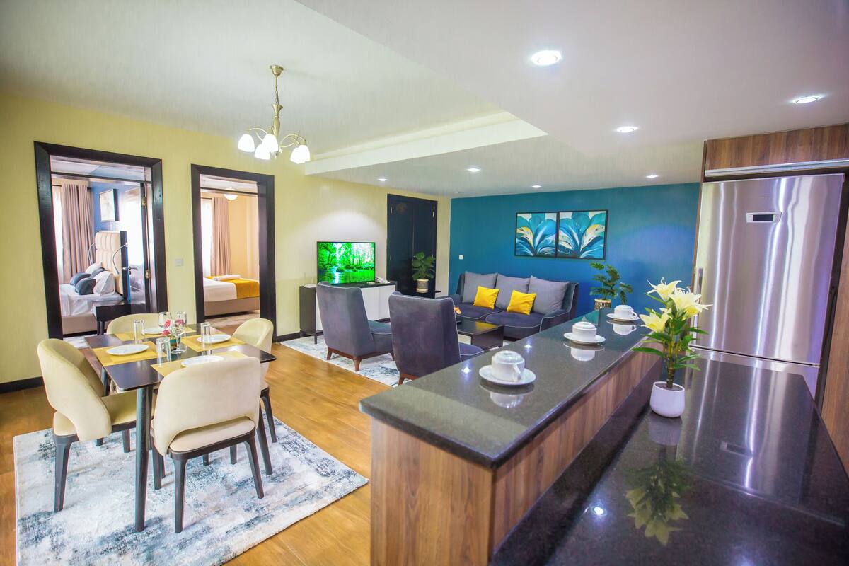 A modern living room with a dining area, a kitchen with a refrigerator, and a TV.