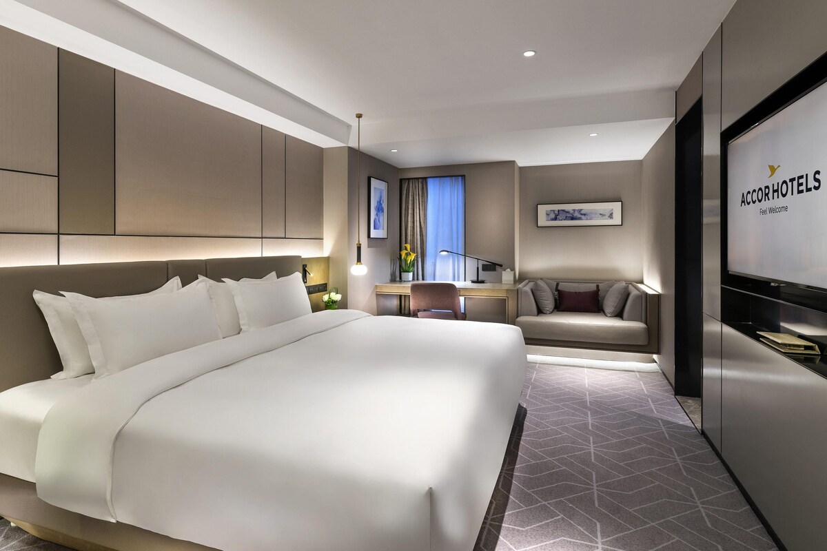 Ein modernes Hotelzimmer mit einem großen Bett, einer Sitzecke, einem Schreibtisch und einem Fernseher, der den Namen des Hotels anzeigt.