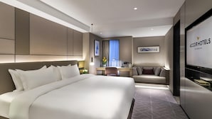 Une chambre d’hôtel moderne équipée d’un grand lit, d’un canapé, d’un bureau et d’un téléviseur affichant le nom de l’hôtel.